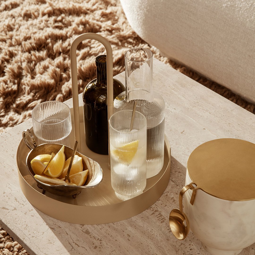 Gold circular tray featuring glassware, a black bottle, and lemon wedges on a textured beige surface. Stylish and modern.