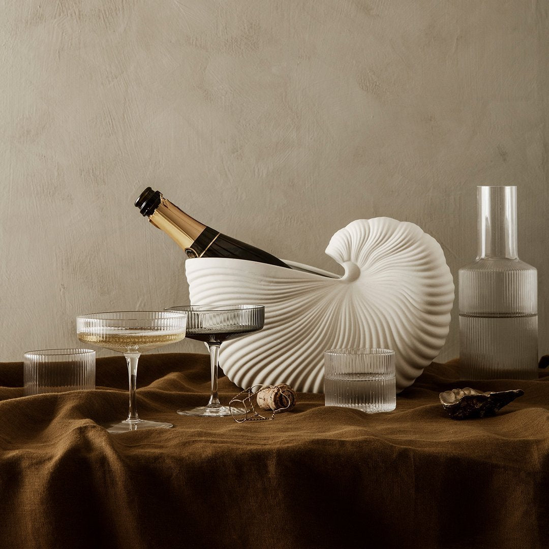 Elegant white ribbed shell vase with champagne, surrounded by clear glassware on a rich brown linen backdrop.