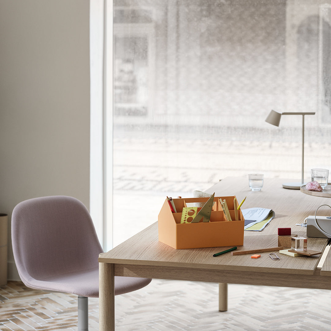 Pastel pink chair and a vibrant orange storage box on a light wood desk; subtle geometric patterns on the floor.